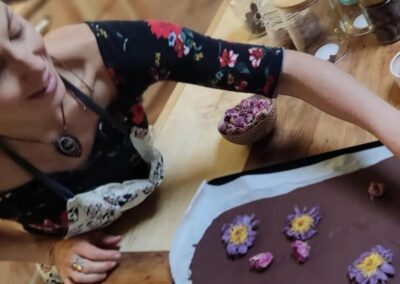 Femme décorant du chocolat avec des fleurs de lotus et des pétales de rose sur une table de cuisine remplie d’ingrédients naturels.